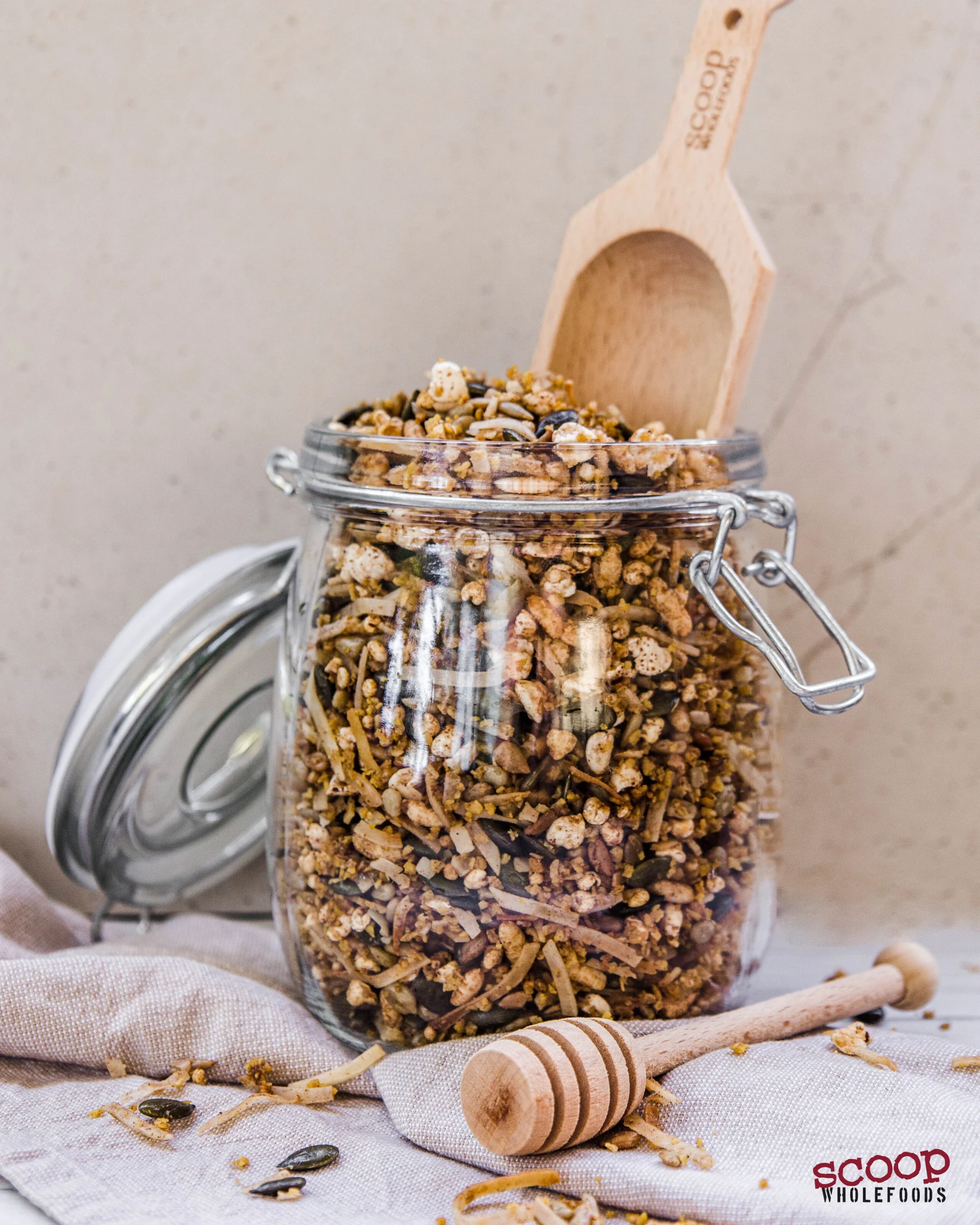 A reusable glass jar of granola with a Scoop spoon, kitchen linen and a honey dipper
